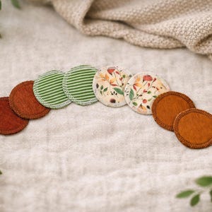 Könnte beinhalten: Eine Sammlung wiederverwendbarer, kreisförmiger Abschminkpads. Einige sind rostbraun, während andere grüne Streifen oder florale Muster in Herbstfarben aufweisen. Die Pads sind auf einer neutralen Leinenoberfläche angeordnet.