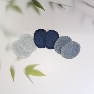 Könnte beinhalten: Eine Sammlung wiederverwendbarer, kreisförmiger Gesichtsreinigungspads in verschiedenen Denim-Blau- und Grautönen. Die Pads sind auf einer weißen Oberfläche angeordnet, mit weichen grünen Blättern im Hintergrund. Diese umweltfreundlichen Pads sind eine nachhaltige Alternative zu Einweg-Wattenpads.