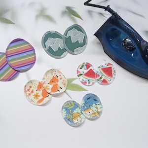 Könnte beinhalten: Sortiment an Bügelbildern aus Stoff mit verschiedenen Designs, darunter Streifen, Füchse, Wassermelonen und Katzen. Die Bügelbilder sind auf einer weißen Oberfläche neben einem blauen Bügeleisen angeordnet.