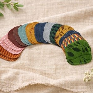 Könnte beinhalten: Eine Sammlung wiederverwendbarer, kreisförmiger Abschminkpads in verschiedenen Farben und Mustern, darunter Streifen, Blätter und florale Designs. Die Pads sind in einem Bogen auf einem neutralen Stoff angeordnet, mit floralen Akzenten.