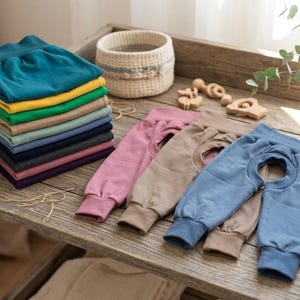Könnte beinhalten: Eine Auswahl an Babyhosen in verschiedenen Farben, darunter Türkis, Gelb, Grün und Rosa. Mehrere Hosenpaare in Rosa, Beige und Blau sind ebenfalls zu sehen. Ein geflochtener Korb und Holzspielzeug sind ebenfalls vorhanden.