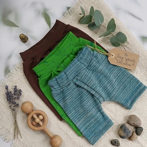 Pu&ograve; includere: Tre paia di pantaloncini per bambini in marrone, verde e verde-azzurro. Il paio verde ha un motivo a foglie. Un'etichetta recita "The Tiny Sprouts Collection". Sono presenti anche un sonaglio di legno, lavanda e piccole pietre.