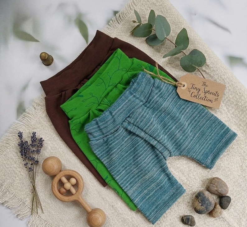 Pu&ograve; includere: Tre paia di pantaloncini per bambini in marrone, verde e verde-azzurro. Il paio verde ha un motivo a foglie. Un'etichetta recita "The Tiny Sprouts Collection". Sono presenti anche un sonaglio di legno, lavanda e piccole pietre.