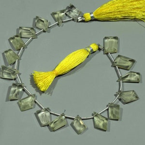May include: A strand of faceted, light green gemstone beads with a silver string. The beads have a geometric shape. Two bright yellow tassels are attached to the ends of the strand. The background is a solid gray.