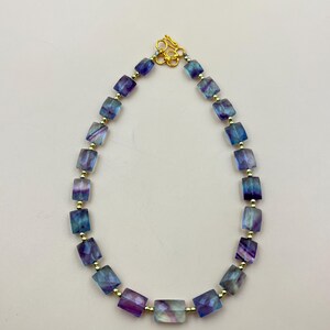Puede incluir: Un collar hecho de cuentas cuadradas iridiscentes azules, moradas y verdes con detalles dorados.