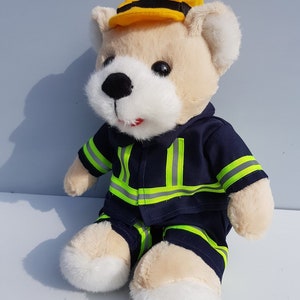 May include: A plush teddy bear wearing a blue and yellow firefighter uniform with reflective stripes. The bear is wearing a yellow hard hat with a black band.