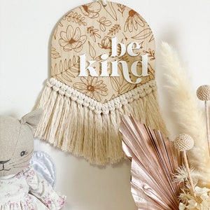 May include: A wooden wall hanging with a floral pattern and the words "be kind" in white letters. The hanging has a fringe of tassels at the bottom.
