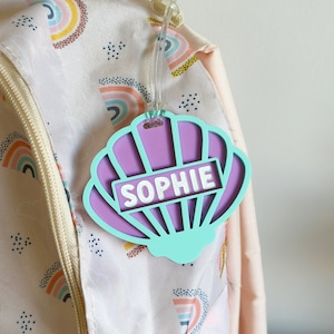 May include: A turquoise seashell-shaped luggage tag with a purple interior and the name "SOPHIE" printed in white.