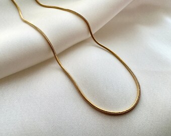 May include: A gold snake chain necklace on a white fabric background.