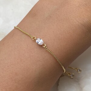 K&ouml;nnte beinhalten: Ein goldenes Kettenarmband mit einem einzigen klaren ovalen Stein in der Mitte. Das Armband hat einen Schiebeverschluss und zwei kleinere klare Steine am Ende.