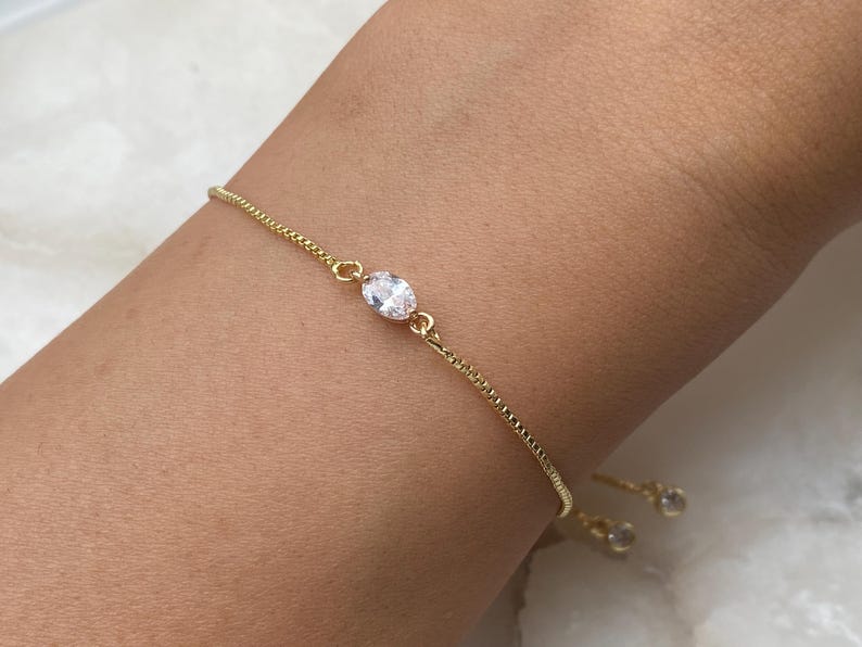 K&ouml;nnte beinhalten: Ein goldenes Kettenarmband mit einem einzigen klaren ovalen Stein in der Mitte. Das Armband hat einen Schiebeverschluss und zwei kleinere klare Steine am Ende.