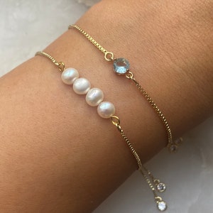 Puede incluir: Dos pulseras de cadena dorada con cierres ajustables. Una pulsera presenta una fila de cuatro perlas blancas y la otra una piedra preciosa azul claro.