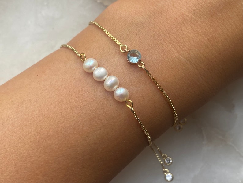 Puede incluir: Dos pulseras de cadena dorada con cierres ajustables. Una pulsera presenta una fila de cuatro perlas blancas y la otra una piedra preciosa azul claro.