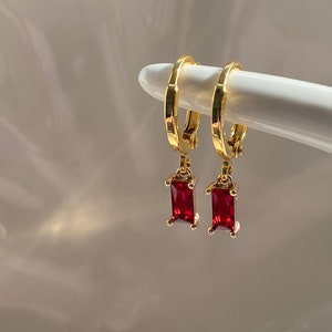 Può includere: Orecchini a cerchio dorati con pietre rettangolari rosse pendenti.