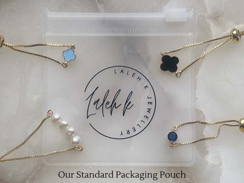 Puede incluir: Cuatro pulseras de cadena dorada con diferentes dijes en una bolsa de pl&aacute;stico transparente. Los dijes incluyen una flor azul, un tr&eacute;bol negro, una fila de perlas blancas y una piedra preciosa azul. La bolsa tiene el texto "Laleh K Jewellery" impreso en ella.