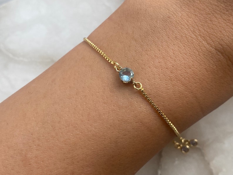 Puede incluir: Un delicado brazalete de cadena dorada con una piedra preciosa azul claro. El brazalete tiene un cierre deslizante ajustable.