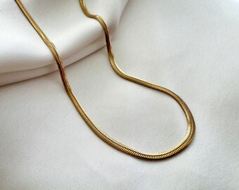 May include: A gold snake chain necklace on a white fabric background.