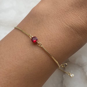 May include: A gold chain bracelet featuring a single red gemstone. The bracelet has a delicate chain and a small, clear gemstone charm.