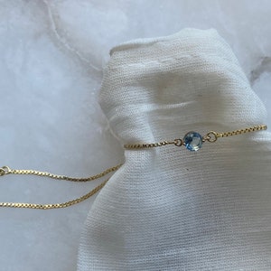 Puede incluir: Un delicado brazalete de cadena dorada con un colgante de piedra preciosa azul claro. El brazalete es ajustable y tiene un cierre deslizante.