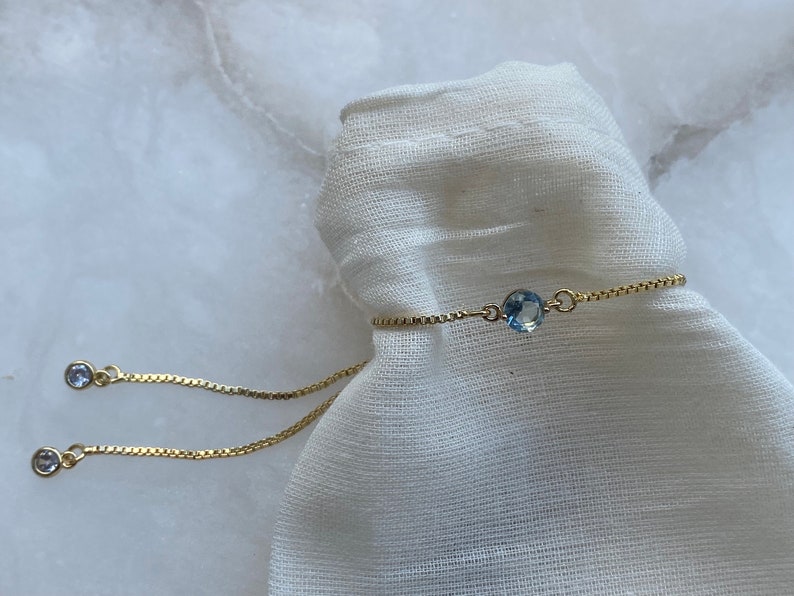 Puede incluir: Un delicado brazalete de cadena dorada con un colgante de piedra preciosa azul claro. El brazalete es ajustable y tiene un cierre deslizante.