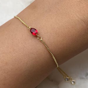 K&ouml;nnte beinhalten: Ein goldenes Kettenarmband mit einem roten ovalen Edelstein. Das Armband hat einen verstellbaren Schiebeverschluss.