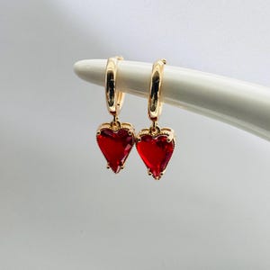 May include: A pair of gold hoop earrings with dangling red heart-shaped charms. The hearts are set with red cubic zirconia stones.