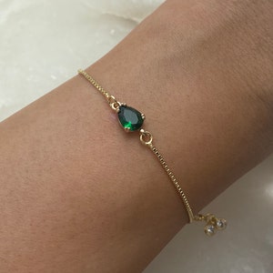 May include: A gold chain bracelet with a green teardrop-shaped gemstone pendant and two small clear gemstones at the end of the chain.