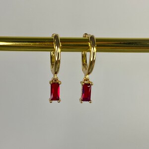 Peut inclure: Boucles d'oreilles créoles dorées avec une pierre rectangulaire rouge pendante.