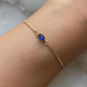 K&ouml;nnte beinhalten: Ein zartes Armband aus einer goldenen Kette mit einem einzigen blauen ovalen Edelstein.