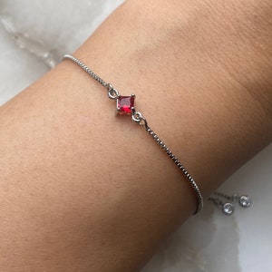 May include: A silver chain bracelet with a red square-shaped gemstone. The bracelet has an adjustable sliding clasp.