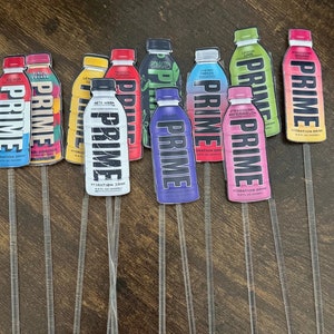 Puede incluir: Un conjunto de diez botellas de bebida de hidratación PRIME de colores vivos con diferentes sabores. Las botellas están dispuestas en fila sobre una superficie de madera. Cada botella tiene un diseño único y el nombre del sabor está impreso en la etiqueta. Las botellas son todas de 12 fl oz (355 ml).