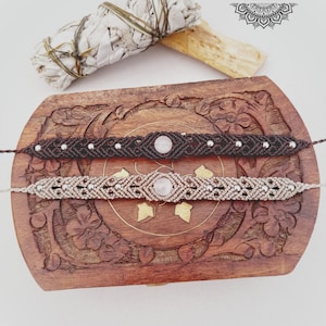 Puede incluir: Dos pulseras de macramé con cuentas de piedras preciosas. Las pulseras son marrones y beige con cuentas plateadas y se muestran en una caja de madera con intrincados tallados. La caja está cerrada y las pulseras están encima.