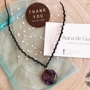 Peut inclure: Un collier en macramé noir avec des perles argentées et un pendentif en forme de rose en améthyste sculptée. Le collier est emballé dans un sac en organza turquoise avec une étiquette de remerciement et une carte de visite qui indique "Aura de Gaia Joyeria en Macrame".