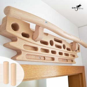 May include: A light-colored wooden hangboard for climbing training featuring various grip holes and slots, mounted above a door frame. The board includes a curved pull-up bar and the text GRIPNATIC. Two small wooden dowels are shown in the corner.