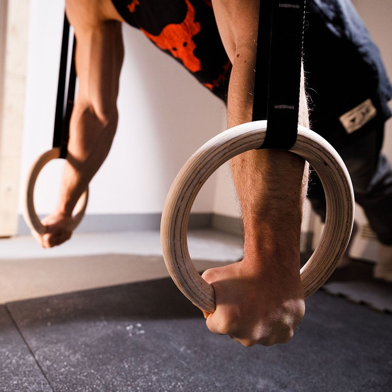 May include: A person working out on gymnastic rings. The rings are made of wood and have black straps.