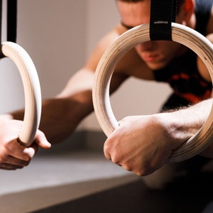 Op de afbeelding: Een persoon houdt twee houten gymnastiekringen vast. De ringen hangen aan zwarte banden aan het plafond.