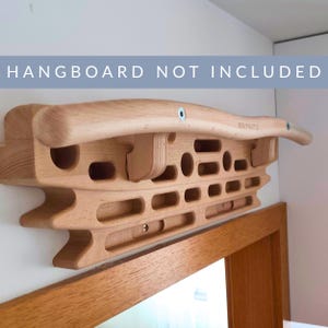 Peut inclure: Un pan de bois pour l'entraînement à l'escalade, avec différentes prises. Le pan est fixé au mur et comporte des poches, des réglettes et des plats. Le texte "HANGBOARD NOT INCLUDED" est visible au-dessus du pan.