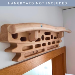 May include: A wooden hangboard for climbing training, featuring a variety of grips such as rounded edges and finger pockets. The hangboard is mounted on a white wall. The text "HANGBOARD NOT INCLUDED" is visible. It is made of light-coloured wood.