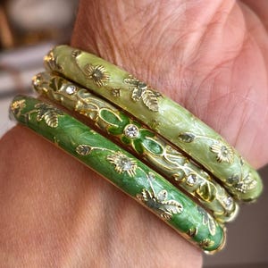 May include: A set of three stacked bangle bracelets in varying shades of green. Each bracelet features a smooth enamel finish with gold-tone metal floral and leaf motifs, accented by small, clear, round inset crystals throughout the designs.