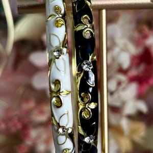 May include: Two circular bangle bracelets, one in white and one in black, both featuring gold-tone floral patterns and embedded clear crystals. The bracelets are displayed side-by-side, showcasing a smooth enamel finish and metallic gold accents.