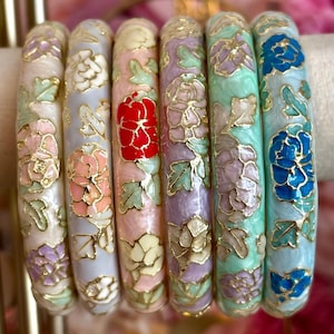 May include: A set of six round bangle bracelets featuring gold-tone metal outlines filled with colourful enamel floral patterns. Each bracelet displays a unique colour palette including shades of cream, lavender, pink, mint green, and light blue.