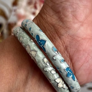May include: Two round bangle bracelets featuring a pale mint green enamel base with a metallic silver finish. The surface is decorated with raised silver floral and vine patterns, accented with small inlaid petals in shades of bright blue and cream.