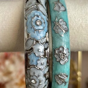 May include: Two metal bangles featuring enamel details. The left bangle displays light blue and white floral patterns with small embedded clear stones. The right bangle is a solid turquoise colour with silver-toned butterfly and flower motifs.