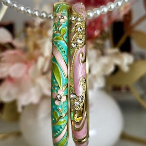 May include: Two gold-tone metal bangle bracelets featuring enamel inlay in shades of turquoise, light pink, and lime green. The bracelets are decorated with floral and swirling patterns and are accented with small, clear, round-cut rhinestones.