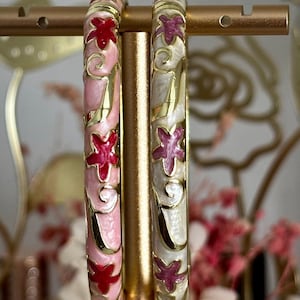 May include: Two circular bangles featuring gold-tone metal edges and enamel inlay. One bangle has a light pink base with red star-shaped motifs and gold swirls. The other bangle has an off-white base with dark pink star-shaped motifs and gold swirls.
