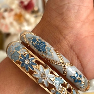 May include: Two gold-tone metal bangles featuring light blue and white enamel floral patterns. One bangle has a solid band with an X-shaped gold detail, while the other features a cut-out design with small clear crystals set in the centre of flowers.