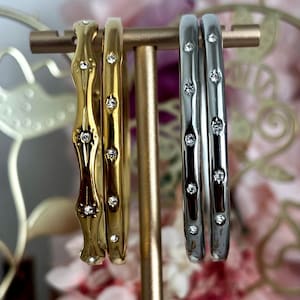 May include: Two gold-toned bangles and two silver-toned bangles displayed on a stand. The gold bangles feature a wavy edge and a smooth band, both accented with small, clear, round-cut stones. The silver bangles have a smooth, rounded profile.