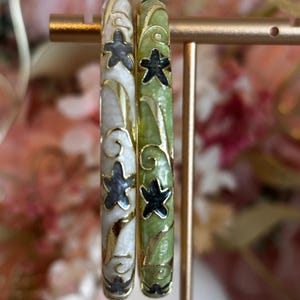 May include: Two circular bangles featuring gold-tone metal accents. One bangle has a white enamel finish with dark grey star motifs, while the other has a light green enamel finish with black star motifs. Both display swirling gold-tone patterns.