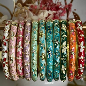 May include: A collection of ten thin metal bangles arranged in a row, each featuring a distinct enamel colour including white, pink, lavender, mint, turquoise, blue, green, orange, and red, decorated with gold floral filigree and small crystals.