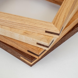 May include: A stack of four wooden picture frames in varying shades of brown and tan. Each frame has a mitered corner with a dark wood accent. The frames are made of different types of wood, showcasing natural grain patterns.